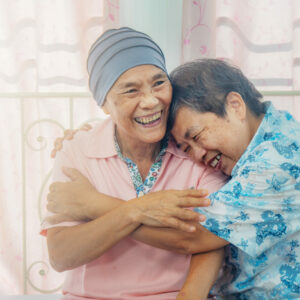 Image of two older Asian ladies sitting on the bed, with one wearing a light blue beanie to cover hair loss form treatment. Her friend is giving her a hug and resting head on her shoulder.